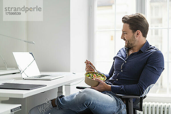 Geschäftsmann mit Salatschüssel und Laptop am Schreibtisch im Büro
