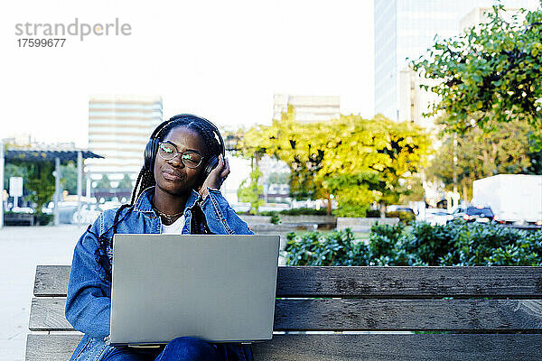 Young woman smiling while sitting on city bench with laptop and headphones