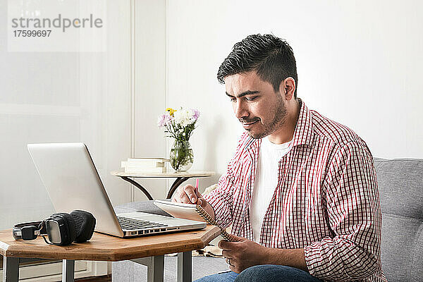 Lächelnder Mann schreibt im Wohnzimmer am Laptop in Notizblock