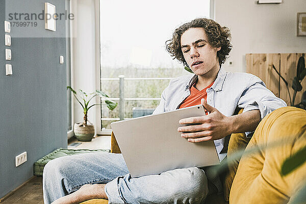 Junger Mann mit braunen Haaren sitzt zu Hause auf dem Sofa und benutzt Laptop