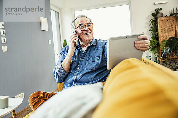 Glücklicher älterer Mann hält Tablet-PC in der Hand und spricht mit Smartphone im Wohnzimmer