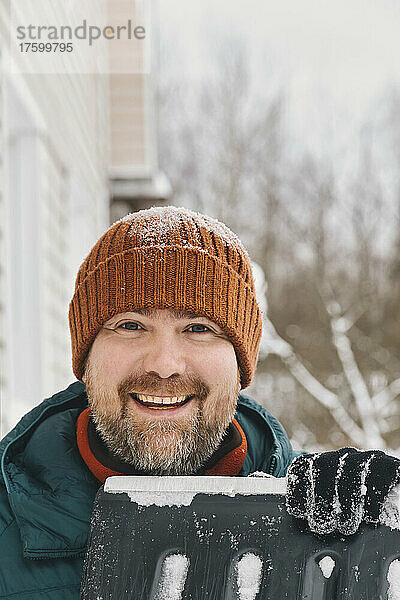 Glücklicher Mann mit Strickmütze und Schneeschaufel im Winter