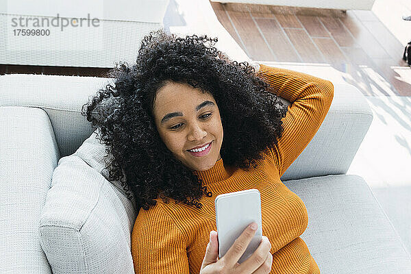 Lächelnde Frau  die auf dem Sofa liegt und ihr Mobiltelefon benutzt