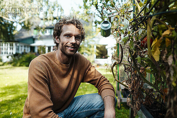 Lächelnder junger Mann kniet neben Pflanzen im Garten