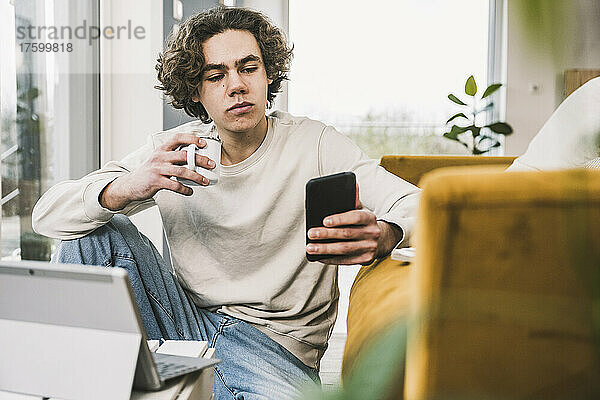 Junger Mann hält Kaffeetasse und Smartphone im Wohnzimmer