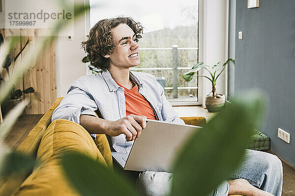 Nachdenklich lächelnder Mann mit Laptop sitzt zu Hause auf dem Sofa im Wohnzimmer