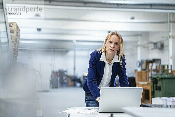 Nachdenkliche blonde Geschäftsfrau mit Laptop lehnt am Schreibtisch in der Fabrik