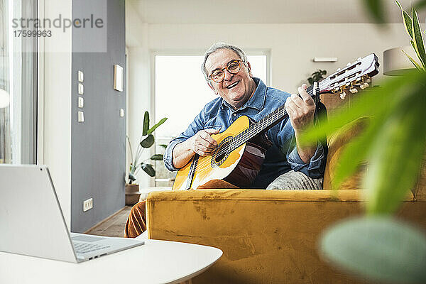 Glücklicher älterer Mann mit Gitarre  der zu Hause auf dem Sofa im Wohnzimmer sitzt