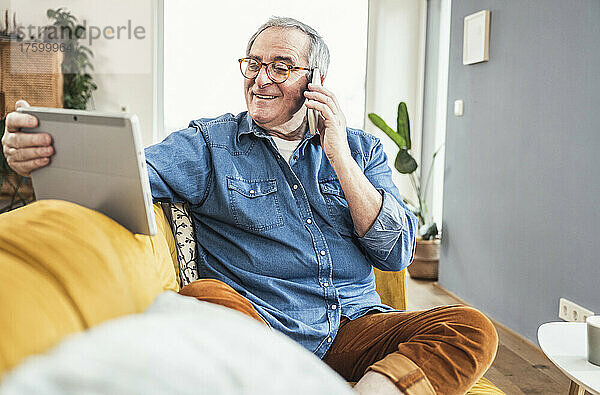 Lächelnder älterer Mann  der mit dem Handy spricht und auf den Tablet-PC im Wohnzimmer schaut