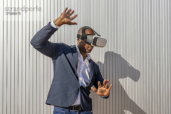 Geschäftsmann mit Virtual-Reality-Simulator gestikuliert an einem sonnigen Tag vor der Wand