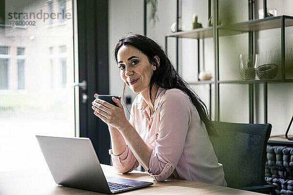 Lächelnde Geschäftsfrau mit Laptop beim Kaffee am Schreibtisch im Coworking-Büro