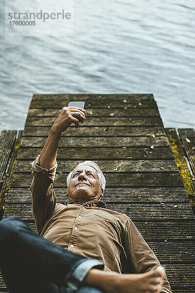 Älterer Mann macht Selfie mit Smartphone und liegt auf Steg am See