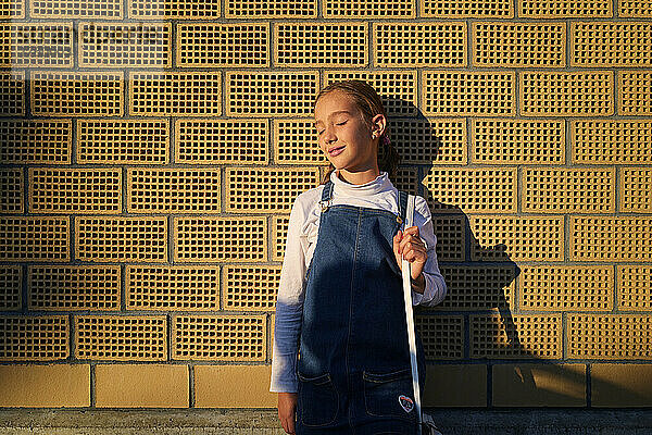 Mädchen mit geschlossenen Augen steht bei Sonnenuntergang vor einer Ziegelmauer