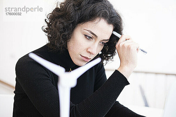Nachdenkliche junge Geschäftsfrau hält Stift neben Windturbinenmodell im Heimbüro