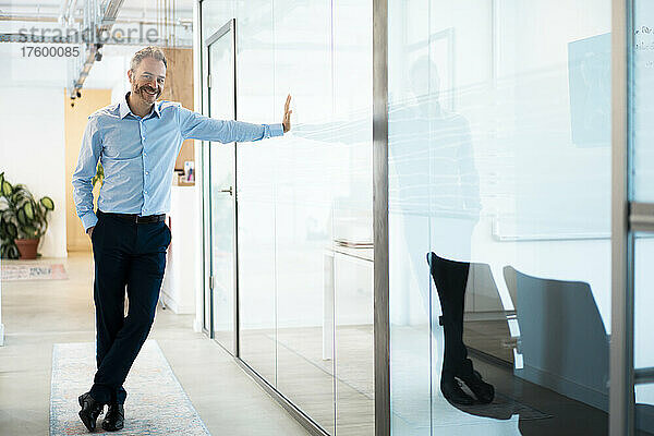 Lächelnder Geschäftsmann lehnt an einer Glaswand im Flur am Arbeitsplatz