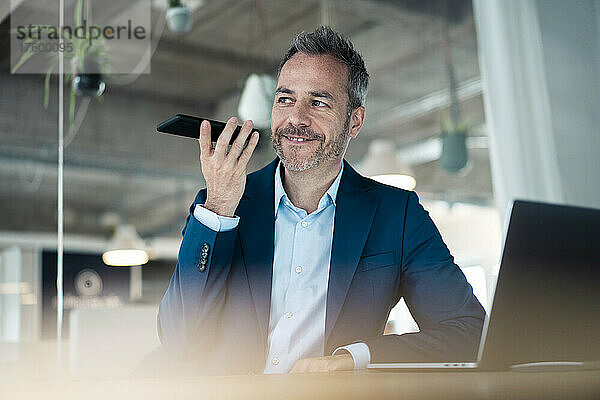 Smiling businessman talking on mobile phone sitting with laptop in workplace