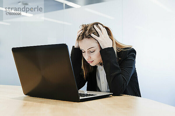 Erschöpfte Geschäftsfrau mit Kopf in den Händen sitzt mit Laptop am Tisch im Büro