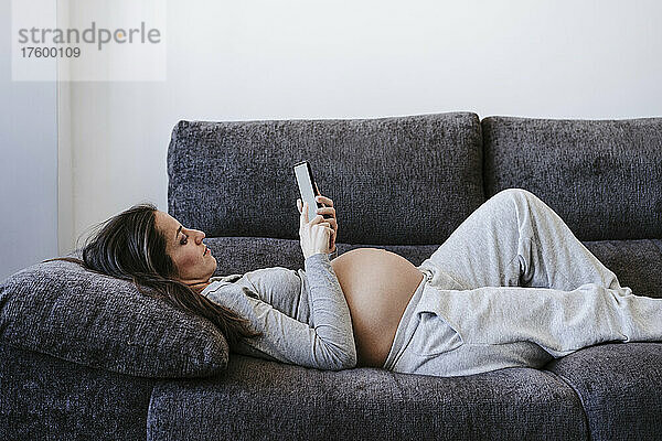 Pregnant woman using smart phone lying on sofa at home