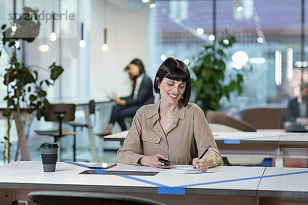 Lächelnde Geschäftsfrau schreibt am Tisch im Büro auf Papier