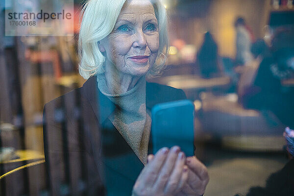 Ältere Frau mit Smartphone durch Fenster gesehen