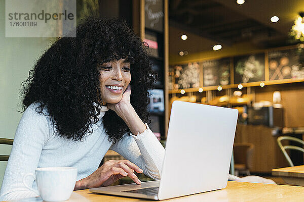 Lächelnde junge Frau mit der Hand am Kinn und Laptop im Café