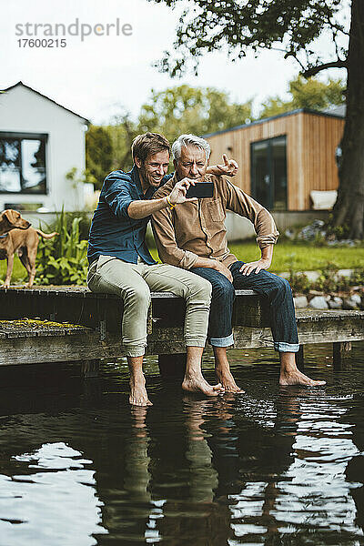 Lächelnder Mann macht mit seinem Vater ein Selfie per Smartphone auf dem Steg am See