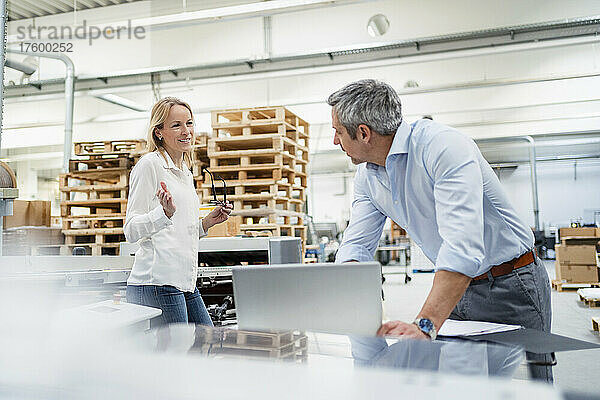Lächelnde blonde Geschäftsfrau tauscht Ideen mit Kollegin aus  die am Schreibtisch in der Fabrik lehnt