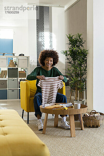 Lächelnde junge Afro-Frau  die ein gestreiftes T-Shirt faltet  sitzt zu Hause auf einem Stuhl