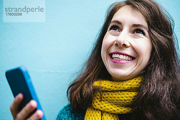 Cheerful woman with smart phone in front of blue wall