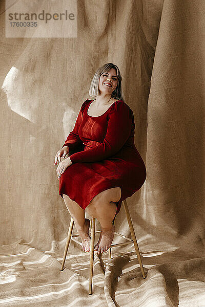 Smiling curvy woman wearing red dress sitting on stool in front of brown background