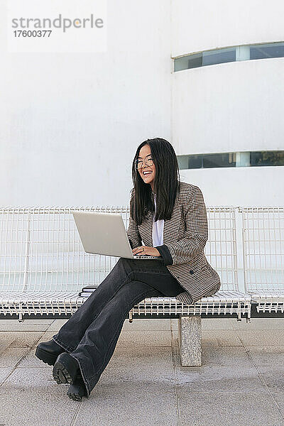 Lächelnde Geschäftsfrau mit Laptop sitzt auf Bank