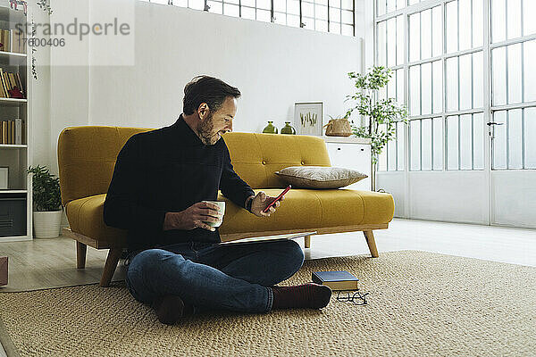 Reifer Mann mit Kaffeetasse und Smartphone im heimischen Wohnzimmer