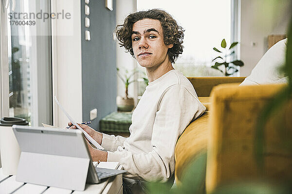 Junger Mann mit Papier und Tablet-PC sitzt zu Hause im Wohnzimmer