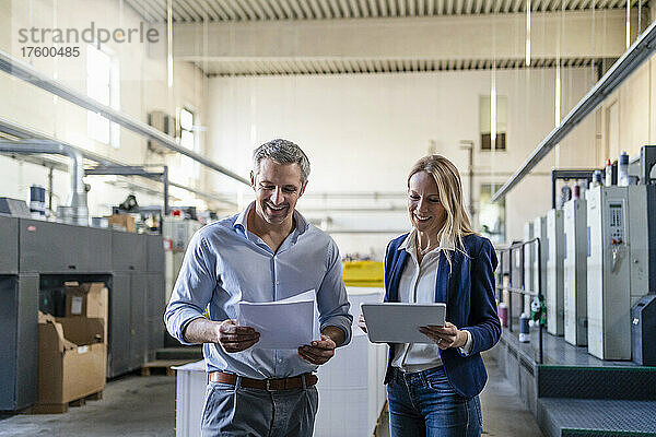 Lächelnde blonde Geschäftsfrau mit Tablet-PC steht neben einem Kollegen und liest Dokument in der Fabrik