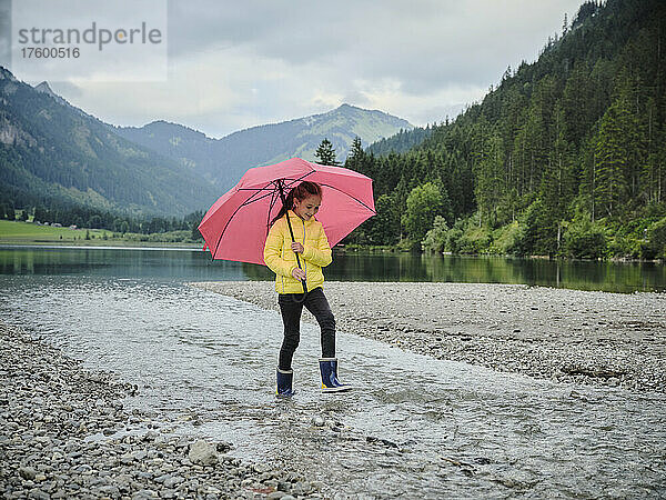 Verspieltes Mädchen mit Regenschirm  das am Seeufer auf dem Wasser spaziert