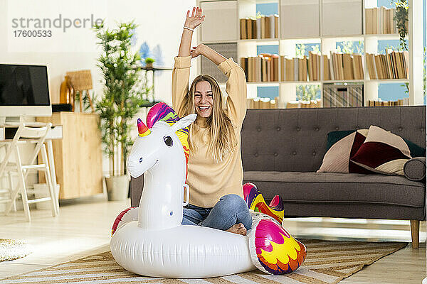 Cheerful woman with arms raised sitting on inflatable unicorn at home