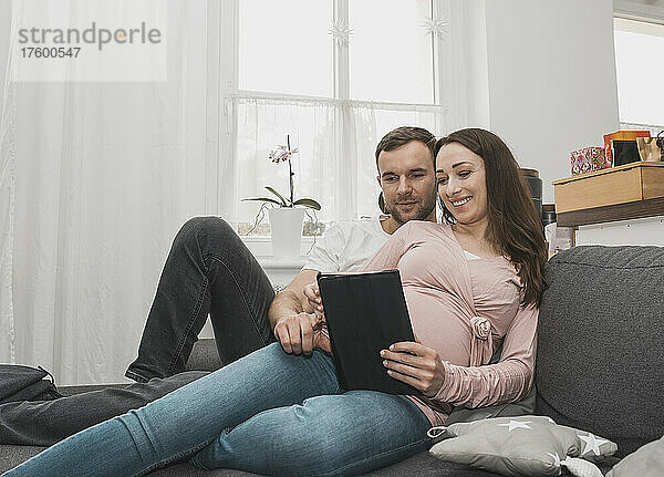 Lächelnde schwangere Frau teilt Tablet-PC mit Mann  der zu Hause auf dem Sofa liegt