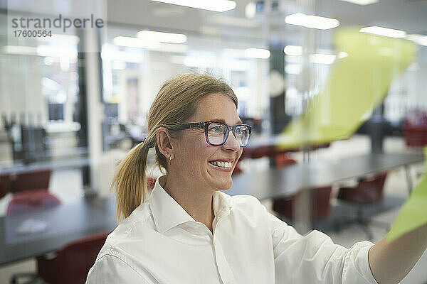 Smiling businesswoman looking at adhesive note on glass wall in office