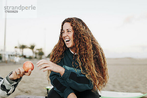 Junge Frau lacht über Freundin  die am Strand ihren Apfel gibt