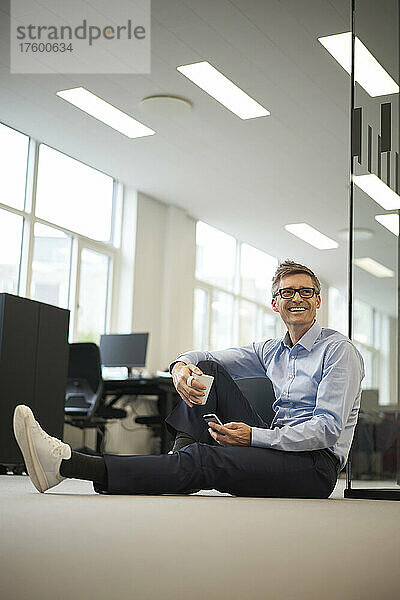 Lächelnder Geschäftsmann mit Smartphone und Kaffeetasse im Büro