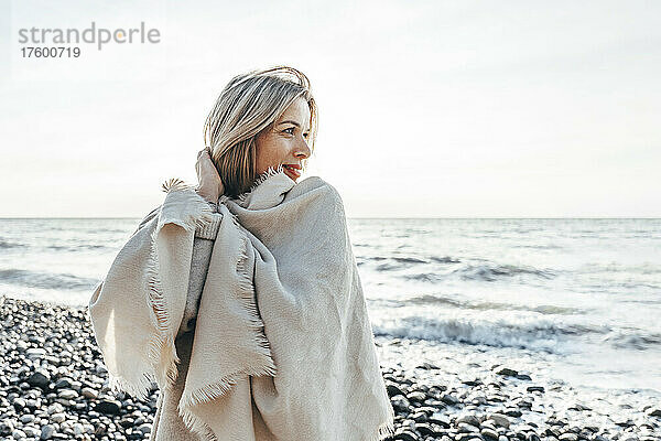 Lächelnde blonde Frau  in eine Decke gehüllt  am Strand