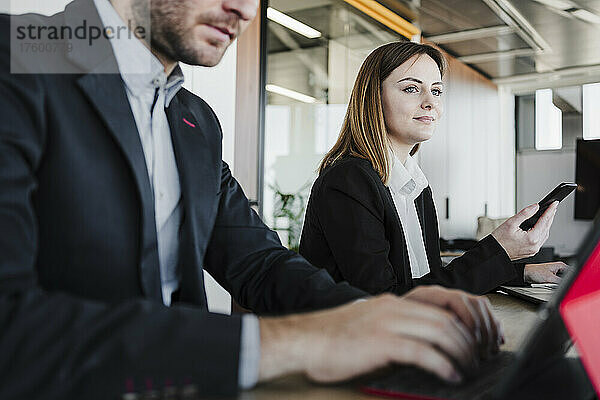 Nachdenkliche Geschäftsfrau mit Smartphone sitzt neben einem Kollegen im Coworking-Büro