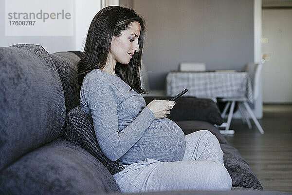Schwangere Frau sitzt zu Hause auf dem Sofa und nutzt ihr Smartphone