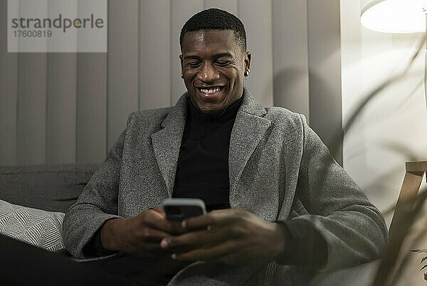 Smiling young man using smart phone sitting on sofa at home