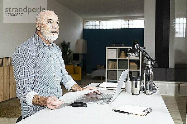 Nachdenklicher Geschäftsmann mit Laptop und Dokument sitzt am Schreibtisch im Heimbüro