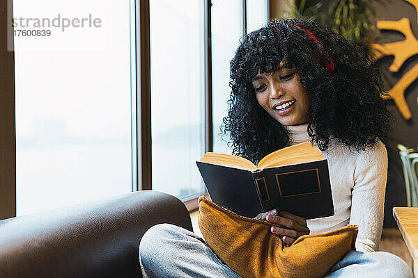 Smiling young woman reading novel sitting on sofa at cafe