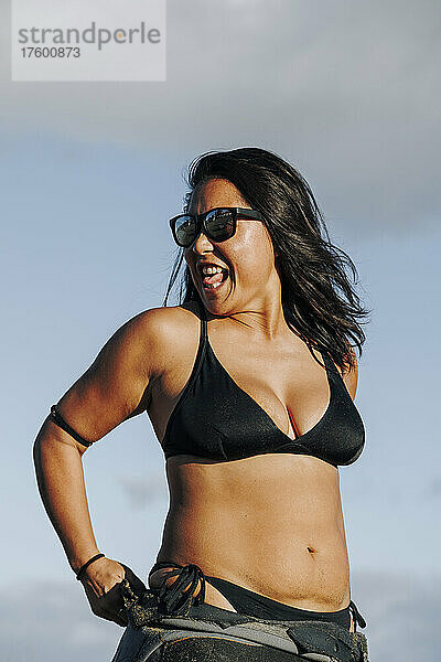 Fröhliche Frau mit Sonnenbrille  Neoprenanzug und Bikini am Strand