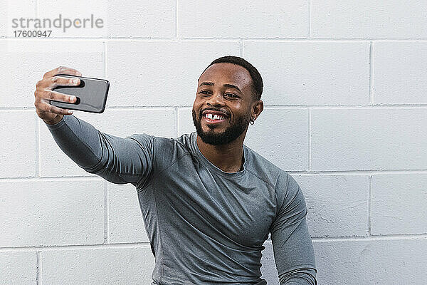 Lächelnder Sportler mit Zahnlücke macht ein Selfie mit dem Smartphone vor der Wand