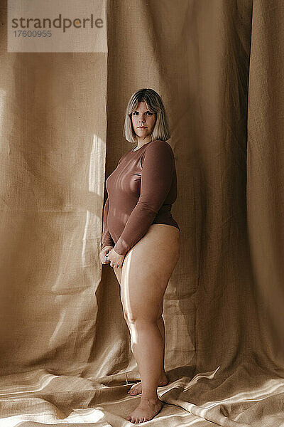 Young curvy woman wearing brown bodysuit standing in front of backdrop