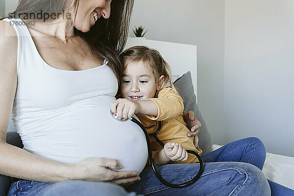 Lächelnde schwangere Frau blickt Tochter an  die durch Stethoskop zuhört